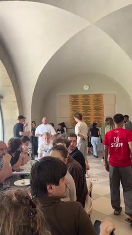 This video shows a gathering of people at tables inside a building with arched ceilings and large windows. The scene appears to be a casual event, possibly a meal or a break, with individuals seated and standing.

In the foreground, several people are seated at a long table, some looking at their phones or eating. The person in the furthest foreground has long, brown, curly hair. To their right, a person with dark hair looks towards the camera with a curious expression. Further down the table, people are engaged in conversation and eating.

In the background, more people are standing and milling around. One person in a red shirt with "STAFF" written on the back is visible. Through the large windows, a bright red food truck with the text "PIZZALEGENDE.CH" is visible, suggesting an outdoor food vendor might be present. The lighting suggests it is daytime, with natural light streaming in from the windows. The overall atmosphere seems relaxed and social.

The architecture of the building, with its stone arches and vaulted ceiling, gives it a historic or institutional feel. The tiled floor further adds to this impression. The presence of people of various ages and ethnicities indicates a diverse group.