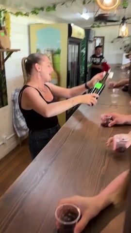 A blonde woman with her hair in a ponytail, wearing a black tank top and blue jeans, is actively pouring red liquid from a green bottle into small plastic cups across a wooden counter. She is speaking and gesturing while serving. Several people, whose hands are visible, are holding cups on the other side of the counter, seemingly engaged in a tasting. In the background, behind the woman, a man in a black t-shirt with "TIGH" (likely part of "FIGHT") printed on it stands near a large refrigerator with a "Майкоп" (Maykop) beer advertisement. The interior features white paneled walls, artificial ivy decorations, and hanging light fixtures with exposed bulbs. A plaid shirt hangs on a hook on the wall. The woman is speaking Russian, aligning with the provided location context of Rassvet, Russia.