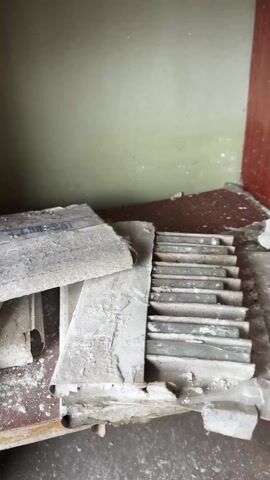 The video reveals a collection of objects in what appears to be an indoor storage area. The backdrop is a light green wall with visible imperfections. In the foreground, there's a cluttered arrangement on a red and brown surface. Various dusty, gray objects are scattered, including what appears to be pieces of broken material and a cardboard container. A hand enters the frame to lift the lid of the cardboard container, revealing a collection of glass ampules nested inside. The ampules appear unused and are arranged in rows. The overall scene suggests a neglected or abandoned space with remnants of storage items.