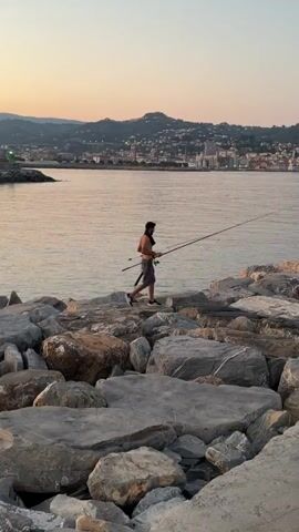 A person is walking along a rocky shoreline, holding two fishing rods. The person is shirtless and wearing shorts and sneakers. The sun is setting, casting a warm glow over the scene. In the background, a city is visible along the coast, with lights beginning to twinkle as dusk settles. The water is calm, reflecting the soft colors of the sky. The overall atmosphere is peaceful and serene. The scene appears to be set in Imperia, Italy, as indicated by the city's characteristic architecture and coastal landscape. No text is visible in the media file.