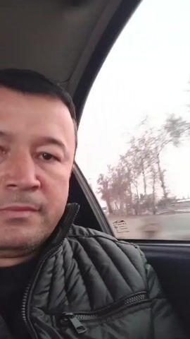 A man is in a car that is moving. The window shows a blurry view of a wintery landscape, including bare trees. The man has short, dark hair and is wearing a black quilted jacket. He is looking at the camera. There is music playing.