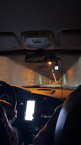 The video shows the interior of a car driving through a tunnel. The camera is positioned from the perspective of the passenger seat, looking forward. The driver's hands are visible on the steering wheel. A smartphone is mounted on the dashboard, displaying what appears to be a navigation app. The tunnel walls are rough and lit by overhead lights, creating a strong contrast between light and shadow. The overall atmosphere is dark and enclosed.