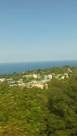 This video captures a scenic drive along a coastal road in Neos Panteleimonas, Greece. The bright blue sky and the shimmering expanse of the sea suggest a clear, sunny day. Lush green trees and vegetation line the roadside, creating a sense of natural beauty and tranquility. In the distance, glimpses of buildings and structures suggest a coastal town or village, adding a human element to the natural landscape. The movement of the camera implies that the footage was recorded from a moving vehicle, perhaps a car or a bus, as it travels along the winding road. The overall atmosphere is one of peaceful exploration and enjoyment of the picturesque surroundings.