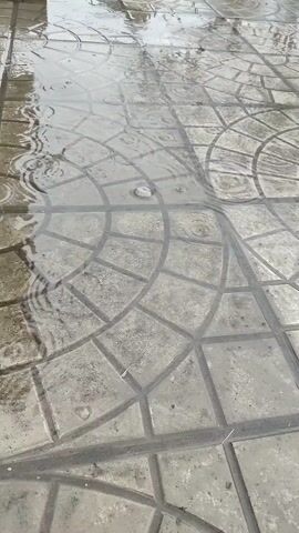 A close-up shot depicts a paved area covered in rainwater. The pavement is composed of gray tiles with a circular pattern design. Raindrops create ripples in the water, distorting the pattern beneath. Green grass is visible in the background.
