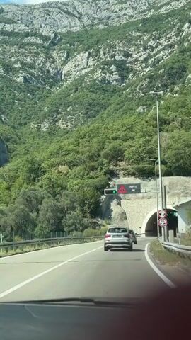 The video shows a car driving on a road towards a tunnel entrance. On the left side of the road, there is a green traffic light and a digital sign displaying a traffic jam icon and text. As the car approaches the tunnel, a red cross symbol appears on a traffic light above the tunnel entrance, indicating closure. Further into the tunnel, a green light and a speed limit sign of 80 are visible. The tunnel is built into a rocky, tree-covered mountainside.