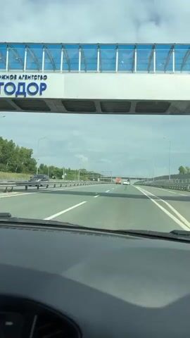 The video shows a first-person perspective of driving on a highway. The road has multiple lanes, with white lines marking the lanes. There are other vehicles visible in the distance, including what appears to be a truck or large vehicle. The sky is cloudy, and there are trees and greenery visible on the sides of the highway, suggesting a rural or suburban environment. Overhead structures like bridges or overpasses are also present. The dashboard of the car is visible in the foreground.