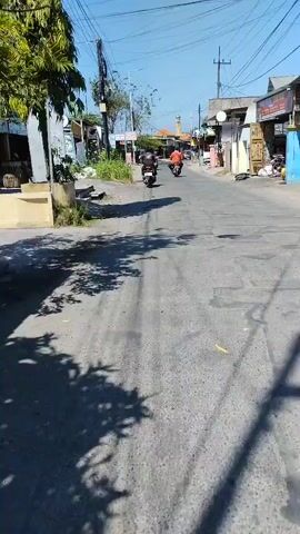 The video shows a street scene, likely in a village or small town, with several motorcycles visible. The road is paved and has some visible damage. Buildings line both sides of the street, appearing to be shops or small businesses. In the foreground, a shadow from trees falls across the road. Several motorcycles are seen on the road, some with riders. The sky is clear and blue, indicating a sunny day. The overall impression is of a typical day in a busy, yet somewhat rustic, urban or semi-urban environment.