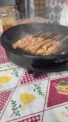 The video shows a person cooking chicken skewers in an electric frying pan. The skewers are laid out in the pan and are being flipped with a fork. The pan is on a table covered with a red and white checkered tablecloth with yellow hearts and teapots. A jar of sauce and a plate of more skewers are visible in the background. The chicken is steaming, indicating it is being cooked at a high temperature.