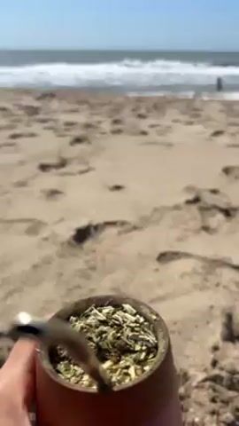 Here's a description of the video:

The video shows a person holding a mate cup filled with yerba mate on a sandy beach. The focus is initially on the mate cup, with a metal straw (bombilla) visible. In the background, the ocean is visible with small waves and a clear blue sky. Several people are standing and playing in the water near the shoreline. The beach is covered in footprints. The camera angle is low, giving a ground-level view.