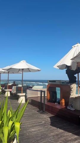 The video shows an outdoor lounge area on a sunny day. In the background, there is a clear blue sky and an ocean. The lounge area has a wooden deck with white umbrellas and cushioned seating. A man wearing a black jacket, tan pants, and a baseball cap is seen adjusting one of the umbrellas. Another person sits in one of the lounge chairs. To the right of the man adjusting the umbrella is a table with audio equipment on it. There are some green plants in the foreground.