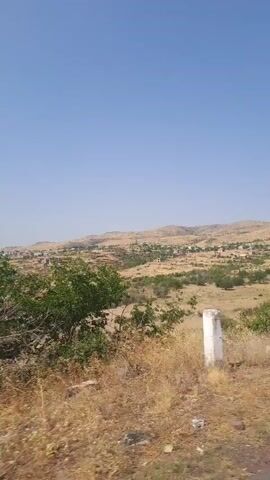 This is a short, vertical video clip, with a duration of 5 seconds. The video showcases a rural landscape under a clear blue sky. The foreground is dominated by dry, brown vegetation and some green bushes. A paved road runs along the bottom right of the frame. In the mid-ground and background, rolling hills are visible, covered in sparse vegetation. A small village or settlement with numerous buildings is scattered across the hills in the distance. The video appears to be shot from a moving vehicle, as indicated by the slight motion blur and the changing perspective. There are no people visible in the video.