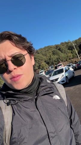 The video shows a person taking a selfie in a parking lot with mountains visible in the background. The person is wearing sunglasses, a black jacket, and a backpack. There are several cars parked in the lot and other people walking around. The sky is clear and blue.