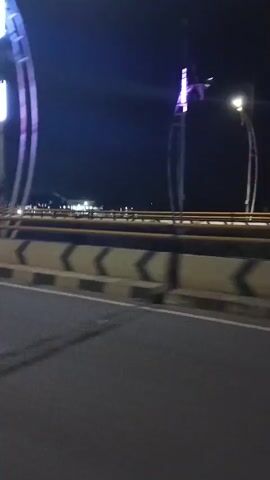 The video shows a nighttime view from a moving vehicle, likely on a bridge. The road surface is dark asphalt. To the right, there is a concrete barrier with black and yellow chevron markings. Beyond the barrier, there are metal railings and lampposts with lights illuminating the scene. In the background, there are structures and more lights, suggesting a city or town. The most prominent features are several tall, arching structures that appear to be decorative streetlights or part of the bridge's architecture, some of which are illuminated with colorful lights (blue, green, and red). The motion blur indicates the vehicle is traveling at a moderate speed. There is no visible interaction between people or any specific actions taking place beyond the movement of the vehicle and the lighting of the structures.