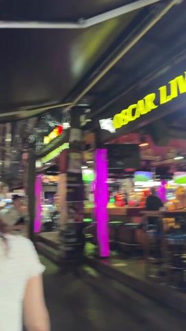 The video begins with a blurred street scene at night, with a couple walking through a crowded area. The scene is illuminated by colorful neon signs, with one sign clearly displaying "TAI PAN CLUB." Various vehicles and pedestrians are visible. The camera then pans, showing a bar called "Oscar" with people sitting on stools, drinking, and watching. The lighting inside the bar is a mix of neon colors, particularly pink and blue. The bar appears busy, with several customers and staff present.