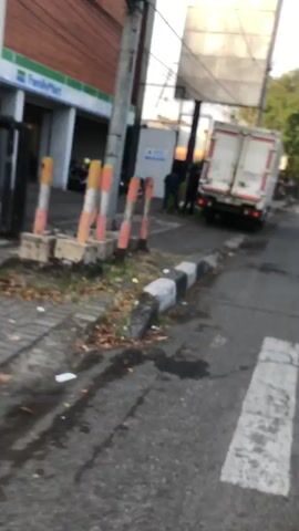The media file captures a street scene in Larangan, East Java, likely during the day given the bright sunlight. A white delivery truck is parked on the right side of the street, its rear doors open. A person, partially obscured, is seen near the back of the truck, interacting with a green container.

In the background, a FamilyMart store is visible on the left, with its distinctive green and white signage. A few motorcycles are parked outside the store, suggesting pedestrian traffic and local commerce. The street is paved and bordered by a curb with distinctive black and white markings. There is some vegetation along the edge of the sidewalk. The overall scene appears to be a typical urban or semi-urban commercial area.

The lighting and shadows indicate it is daytime. No specific weather conditions are evident, but the sunlight suggests it is not raining. The presence of a delivery truck and a convenience store points to a commercial or retail environment. The mood of the scene is neutral, simply documenting a moment in time. Visible text includes "FamilyMart" on the store's facade and "BARANG GRATIS" on a blue sign near the delivery truck.