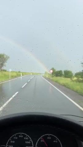 A driver's perspective video shows a wet asphalt road stretching into the distance under a gray, overcast sky with a rainbow arching overhead. Raindrops are visible on the windshield, obscuring the view slightly. White lines mark the road lanes and edges. Green grass and sparse vegetation line the sides of the road. A round, white traffic sign with a red border and horizontal red bar is visible on the right side of the road. The car's dashboard and steering wheel are partially visible at the bottom of the frame. The camera appears to be stationary as the vehicle is in motion, suggesting a dashcam or mounted phone recording.