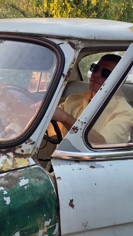 A man wearing sunglasses and a yellow shirt sits in the driver's seat of a white car with peeling green paint. He is holding the steering wheel, and the car appears to be moving, as indicated by the motion blur. The environment seems to be outdoors, with trees visible in the background.