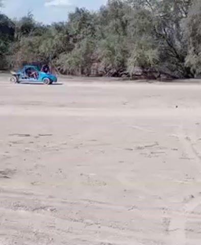 The video shows a light-blue buggy with two occupants sitting on a sandy surface surrounded by trees and bushes. The sky is blue with patches of white clouds. The buggy is still, with visible tire tracks in the sand.