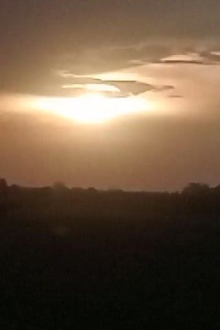 This is a video clip showing a natural landscape at sunset. The sun is a bright, hazy orb low in the sky, casting a warm, golden light through the clouds. The sky is a mix of orange, yellow, and muted gray tones. In the foreground, there is a dark, silhouetted landscape, likely fields or rolling hills, with some indistinct shapes that could be trees or distant buildings along the horizon. The overall mood is peaceful and serene, with the fading light of day. The time of day is clearly sunset. There is no visible text in the video. The location context provided is Pulorayeuk, Indonesia.