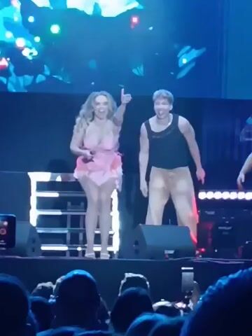 The video shows a woman singing into a microphone on a stage. She is wearing a pink dress and heels. Two men stand near her, appearing to be backup dancers. The stage is set up with speakers and lighting, and there is a large screen behind the performers displaying colorful graphics. An audience is visible in the foreground, watching the performance and recording with their phones.
