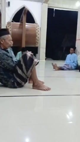 The video shows a group of men sitting on the floor of a building with a tiled floor and concrete pillars. Some of the men are wearing skullcaps. One man in the foreground is wearing a sarong and is sitting with his legs crossed. To his left, a large drum is leaning against a pillar. Further back, several men are seated, facing away from the camera or to the side. One man in the background is wearing a blue shirt and a patterned sarong. Another man is wearing a green shirt and a black bag is over his shoulder. The lighting appears to be artificial, with overhead lights illuminating the space. The background is dark, suggesting it is nighttime or an indoor space with limited natural light. There is a staircase visible in the background.