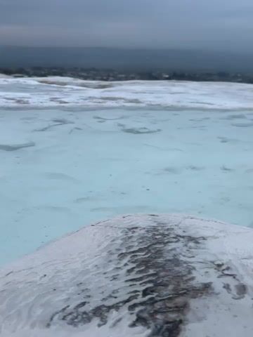 The video shows a body of clear, turquoise water atop a white, textured surface, likely travertine terraces. The water is shallow, revealing the underlying terrain. In the background, a hazy, gray sky contrasts with a distant green landscape dotted with a small settlement of buildings. The perspective is from a slightly elevated position, looking down at the water and out towards the horizon. The general atmosphere is serene, despite the overcast sky.
