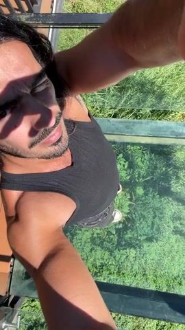 The video shows a man with long dark hair and a beard wearing a black tank top and black jeans. He is standing on a glass floor, looking down at the green grass below. The camera is positioned above him, giving a bird's-eye view. The man appears to be taking a selfie or recording a video of himself. The environment appears to be outdoors on a sunny day.