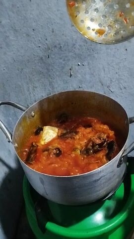 The video shows a silver pot with a red sauce inside, being stirred with a slotted spoon. The pot rests on a green plastic object, and the scene is filmed from above against a grey background. The red sauce seems to contain pieces of fish and something yellow, possibly an egg.
