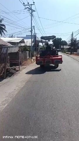 A red tractor with a tilling attachment is driving down a paved road. The tractor has a canopy for shade and a person is driving it. The road is lined with houses and businesses on one side and a dirt shoulder on the other. There are power lines overhead. The sky is clear and the sun is shining. A shadow of the tractor is cast on the road.