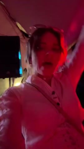 A person is dancing with their head and body partially obscured by a white top and a beige strap across their chest. The scene is illuminated by red, green, and blue lights, with the person's face occasionally visible. They appear to be wearing a pearl necklace and some kind of hair accessory. The background is dark and out of focus.