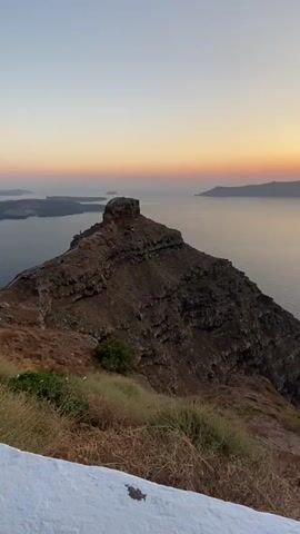 The video opens with a view of a rugged, dark rock formation in the foreground, with sparse vegetation on its slopes. Beyond the rock formation, a vast expanse of calm water stretches to the horizon. In the distance, several islands are visible as silhouetted landmasses against a clear sky. The sun is low in the sky, casting a warm, orange glow that blends into softer pink and yellow hues, indicating either sunrise or sunset. The water reflects the colors of the sky, creating a serene and tranquil atmosphere. The camera pans slightly to the right, revealing more of the water and the distant islands. A small boat or two can be seen on the water in the far distance. The foreground also shows a white, slightly textured surface at the bottom of the frame, suggesting a wall or ledge. The overall scene is a natural landscape with a focus on the dramatic geological formations and the atmospheric lighting over the water.