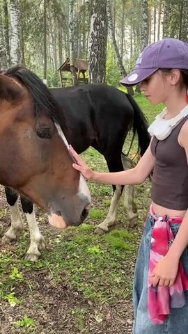 A young person wearing a purple baseball cap, a brown tank top, and blue jeans with a red and pink floral print sash tied around their waist, gently strokes the face of a brown horse. Another black horse with white markings stands behind the brown horse. They are in a wooded area with trees and green grass on the ground. The person has white headphones around their neck.