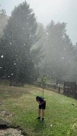 A person wearing a black t-shirt and black shorts is standing in a backyard during a heavy downpour. The person is hunched over with their arms covering their head, seemingly seeking shelter from the rain. The backyard is covered in green grass, and a wooden fence is visible in the background. Several large trees are also present, their leaves obscured by the rain. The sky is overcast and gray, indicating stormy weather.