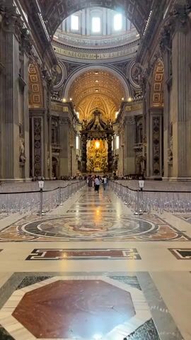 The video captures an indoor, medium shot, eye-level perspective of a majestic, ornately decorated cathedral, likely St. Peter's Basilica, given its grandeur. The camera is fixed, providing a steady view down a long, central aisle. The aisle is flanked on both sides by waist-high, white barriers with black bases, creating a clear path toward the altar. The floor is an impressive mosaic of various colors, predominantly white, brown, and grey, arranged in intricate geometric patterns and medallions. 

In the distance, at the end of the aisle, a magnificent golden altar or canopy, possibly Bernini's Baldacchino, stands beneath a towering dome. Its gilded surfaces reflect the light, creating a focal point. Directly above the altar, a large dome with multiple windows allows natural light to stream into the cathedral, illuminating the space. The ceiling of the nave is equally ornate, featuring a coffered design with gold detailing and recessed lighting fixtures that add to the illumination. 

Numerous columns and pilasters, adorned with sculptures and relief work, line the sides of the aisle, supporting the grand arches and vaulted ceilings. The walls are rich with classical architectural elements, including cornices, friezes, and decorative panels. The overall color scheme is warm, with a predominance of gold, cream, and various shades of marble.

Scattered throughout the visible parts of the aisle and around the altar area are a handful of people, mostly appearing as small figures due to the distance. They are casually walking and observing the cathedral's interior, their actions suggesting they are tourists or visitors. Their attire is indistinct due to the distance. The overall impression is one of awe-inspiring scale, intricate artistry, and historical significance, with the natural light from the dome contributing to the ethereal atmosphere.