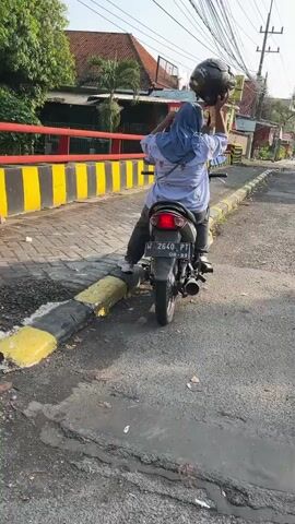 The video shows a person from behind sitting on a motorcycle. The person is wearing a blue hijab and a white shirt. They are holding a black helmet above their head, appearing to be about to put it on. The motorcycle is dark-colored and has a license plate that reads "B 2640 PT." The motorcycle is parked on an asphalt road with yellow and black striped curbing. In the background, there are buildings with red roofs, trees, and power lines. The lighting suggests it is daytime.