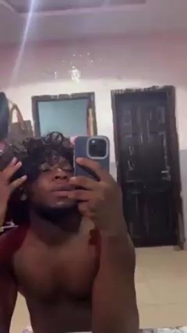 A topless man stands in front of a bathroom mirror, holding a phone up to take a selfie. He has dark skin, curly black hair, and a light beard. He is adjusting his hair with his left hand. The bathroom has a white ceiling and two doors: one black and one smaller, partially mirrored. A purse hangs near the smaller door. The lighting in the bathroom cycles through different colors, including green, red, and blue. The mirror is slightly dirty, with what appear to be water spots.