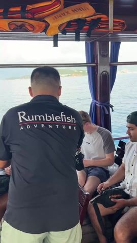 The video shows the inside of a boat with several people standing and sitting. There are life jackets stored in the overhead. A man wearing a black shirt with the words "Rumblefish Adventure" is standing with his back to the camera, followed by two seated passengers. More people appear as the camera pans to the right, including two men wearing black shirts; one has a flag on his sleeve, and the other shirt says "Sailing Club Divers." A red crate sits near them. A woman with a floral dress and a man in a light grey shirt are also standing. Other men in dark shirts are visible near a map and a bag of bananas.