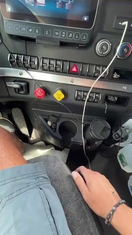 A person's hand, wearing a bracelet, reaches towards the dashboard of a truck. The dashboard has multiple buttons and dials. In the background, there are several bottles of water and Gatorade, along with a paper bag. The person then picks up a large wrench. The scene appears to be inside the cab of a semi-truck.