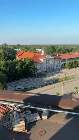 The video captures an elevated, panoramic view of a town or village under a clear blue sky. The camera slowly pans from left to right, revealing different aspects of the settlement.

In the initial frames, a large, ornate building with a light-colored facade, decorative architectural elements, and a prominent red tiled roof is visible in the midground. In front of this building, there is a paved public square or plaza furnished with benches. A road runs along the edge of the square, and a few small, indistinct human figures can be seen near the building. Abundant green trees surround the buildings and extend into the background.

As the camera pans to the right, the large building moves out of frame, and a white church with a red roof, a dome, and a cross on its steeple comes into view in the distance, nestled among the trees. The road continues through the town, and more residential-style buildings are scattered throughout the verdant landscape. The distant horizon appears low and flat, suggesting a wide, open area beyond the town.

In the immediate foreground, a rusty metal railing and some debris, possibly construction materials like broken bricks or concrete, are visible on what appears to be a rooftop or elevated platform. The strong, clear light and long shadows cast by the foreground elements suggest either early morning or late afternoon. The weather is sunny and cloudless. No discernible text is visible.