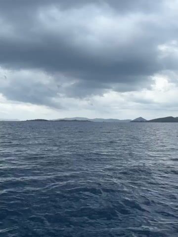 A wide shot shows a choppy, dark blue ocean under a heavily clouded sky. In the distance, a dark, mountainous island chain is visible on the horizon. The foreground is occupied by the front of a white boat with a silver railing. The camera pans slightly to the right, revealing more of the boat's railing and deck. The weather appears overcast and possibly stormy. There are no people visible in the video.