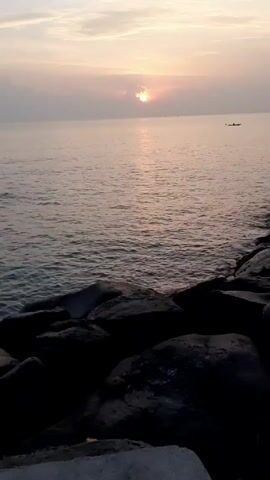 The video shows a seascape at sunset or sunrise. The sun is partially obscured by clouds, casting a shimmering reflection on the calm water. In the distance, a small boat is visible. Large, dark rocks line the shore in the foreground, providing a silhouetted contrast to the soft, pastel colors of the sky and water.