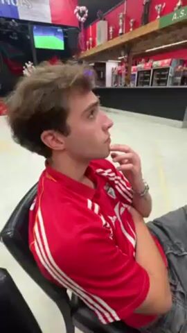 The video shows a young man sitting in a black chair, wearing a red and white soccer jersey. He is resting his chin on his finger, looking pensive. In the background, a television is mounted high on the wall, displaying a soccer game. The room appears to be a gathering place, possibly a club or community center, decorated with sports memorabilia, trophies, and banners. The walls are painted red and white. The man appears to be watching the game on TV.