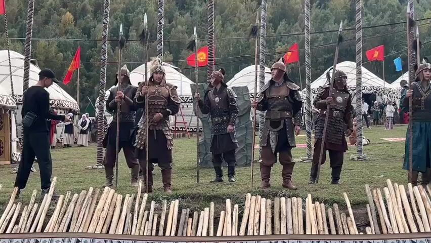 The video captures a historical reenactment or cultural performance in an outdoor setting. In the foreground, a short, rustic wooden fence made of bundled sticks is visible. Behind the fence, a group of approximately seven individuals are standing in formation, dressed in historical armor and holding long spears. Their attire appears to be of Central Asian origin, possibly related to nomadic warriors, featuring leather, metal plates, and elaborate helmets. Some individuals wear turbans or head coverings. The background reveals several large, white yurt-like structures, suggesting a traditional encampment or festival. Behind the yurts, a line of tall wooden poles with flags is erected. The flags are predominantly red with a yellow emblem, likely representing the national flag of Kyrgyzstan. Trees with autumn foliage are scattered across a grassy hillside in the distance, and a mountain range is faintly visible. The lighting suggests it is daytime, possibly late afternoon. One person on the far left, not in costume, is holding a mobile phone, likely recording the event. The atmosphere seems festive and communal, with other people visible in the middle ground near the yurts.