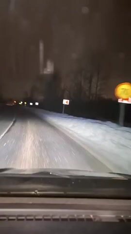 A car drives along a snowy road at night. The road is clear of snow, but the shoulders of the road are covered. Yellow streetlights and road signs are visible in the distance. The car's headlights illuminate the road ahead. The sky is dark and overcast. The vehicle’s dashboard is visible at the bottom of the frame. There are a few water droplets on the windshield.