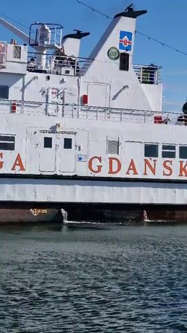 A white ferry with red lettering is visible from the side as it moves through the water. The lettering on the side of the ship reads "ZEGLUGA GDANSK". There are people visible on the upper decks of the ferry, and life preservers are hanging on the side. The water is choppy, and the sky is blue with some clouds. Water is seen being expelled from the side of the ferry near the waterline.