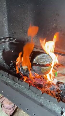 A log is burning in a metal fire pit. The fire is intense, with bright orange flames engulfing the log and the surrounding embers. The log appears to be made of dark wood with visible grain and has a split down the middle. Underneath the log, burning paper and what looks like crumpled aluminum foil can be seen. The fire pit itself is made of metal and has a serrated edge. The background is dark and appears to be the interior of a fireplace or grill. The lighting suggests it is either evening or indoors. There is no visible text in the media.