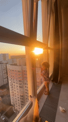Toddler watches sunset at window while adult rests nearby