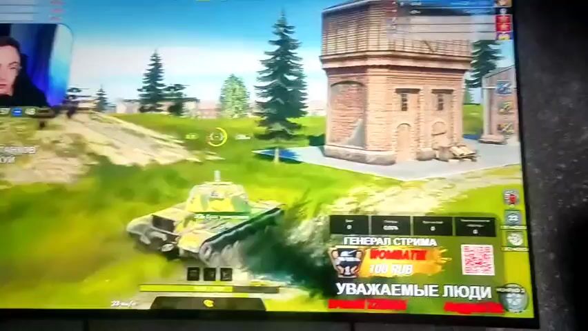 A person is playing a video game on a large screen. The game appears to be a tank battle. The person is looking intently at the screen, with their mouth slightly open. In the foreground of the game, there is a tank. In the background, there are trees and buildings. There is also a small window in the top left corner of the screen showing the person playing the game. There is a donation prompt on the right side of the screen.