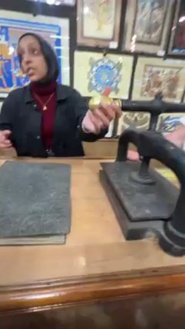 Here's a detailed description of the video:

The video shows a woman in a black jacket and a dark hijab standing behind a wooden counter in what appears to be a store or workshop selling Egyptian artwork. The woman is facing toward the right, gesturing, and talking. In front of her on the counter is a stack of gray, felt-like material next to a black, vintage-looking press with a gold-colored knob on top. The press is centered on the counter.

To the right of the woman stands a person in a white top; only part of their arm is visible. In the background behind the woman, there are numerous framed pictures, presumably artwork for sale, hanging on the wall. The lighting in the space appears bright, with a mix of artificial and possibly natural light. The woman is seen handling the edge of one of the pieces of gray material near the end of the video.