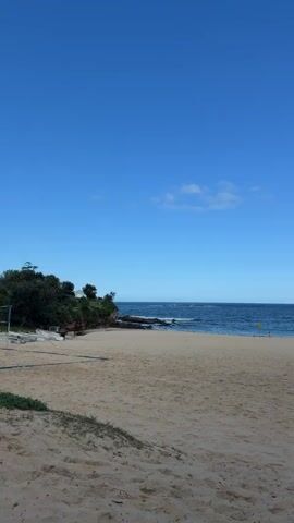 A wide shot of a sandy beach under a clear blue sky. The ocean is visible with gentle waves. On the left side of the frame, there is a rocky outcrop covered in green foliage. The beach is mostly empty, with a single person in the distance using a metal detector. Further down the beach, buildings can be seen on a hill overlooking the ocean. The camera pans slightly to the right, revealing a stone wall and some people walking on it.
